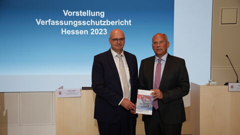 Innenminister Poseck hält Vefassungsschutzbericht in Kamera. Im Hintergrund steht geschrieben: Vorstellung Verfassungsschutzbericht Hessen 2023